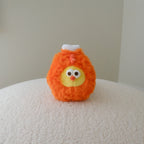 A small orange fluffy drumstick plush toy with a yellow face, black eyes, and a tiny white bone on top, standing on a textured cream surface against a plain light gray wall.