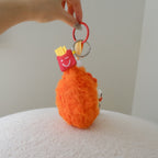 An orange fuzzy drumstick plush keychain with a red ring, held by a hand over a white textured surface. The keychain includes a red french fries bag charm.