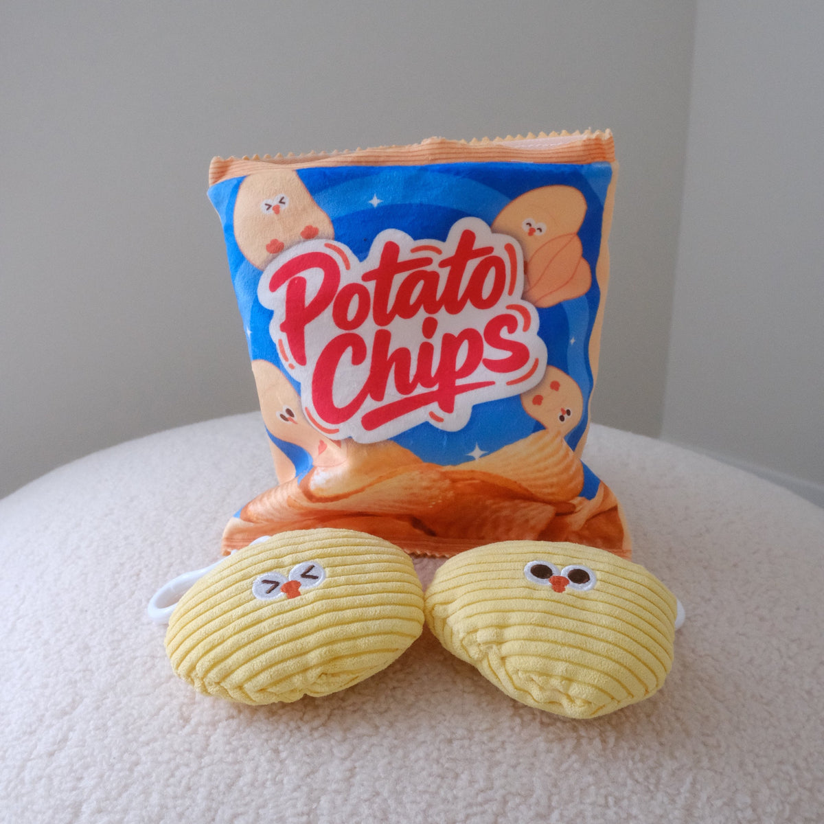 A novelty plush toy set shaped like a bag of potato chips and two potato chip characters, displayed on a soft, light-colored textured surface against a neutral gray background.