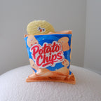 A novelty plush toy shaped like a bag of potato chips sits on a white textured surface against a plain light gray wall. The toy bag is blue with red 'Potato Chips' lettering, orange edges, and cartoon potato chip illustrations.