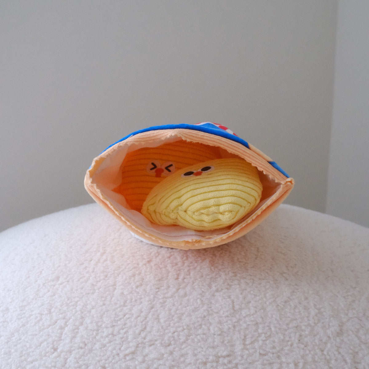 A small plush toy shaped like a clam or shell, shown open on a textured white surface against a plain light gray background. The toy has a peach-orange shell exterior, a yellow coiled body inside, and a blue top edge.