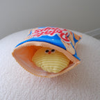 A novelty plush toy shaped like a potato chip bag sitting on a white textured surface against a neutral gray background.