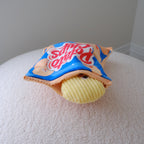 A novelty plush toy shaped like a potato chip bag sitting on a textured white surface against a plain gray wall.