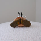 A small plush cockroach keychain with olive green wings resting on a white textured surface against a plain light gray background.