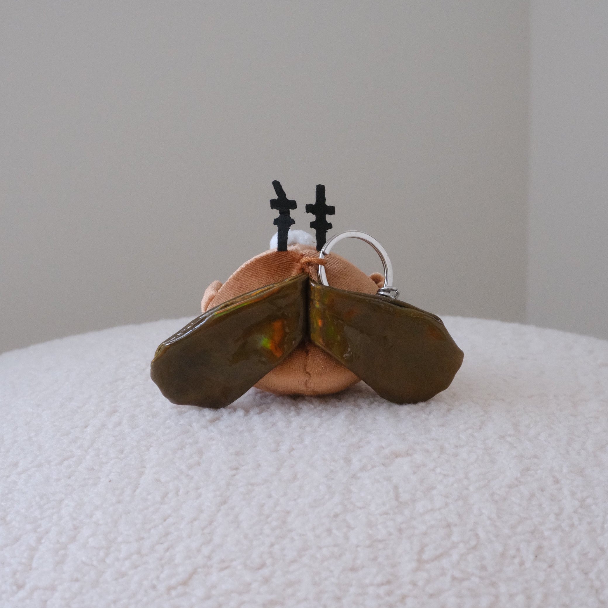 A small plush cockroach keychain with olive green wings resting on a white textured surface against a plain light gray background.