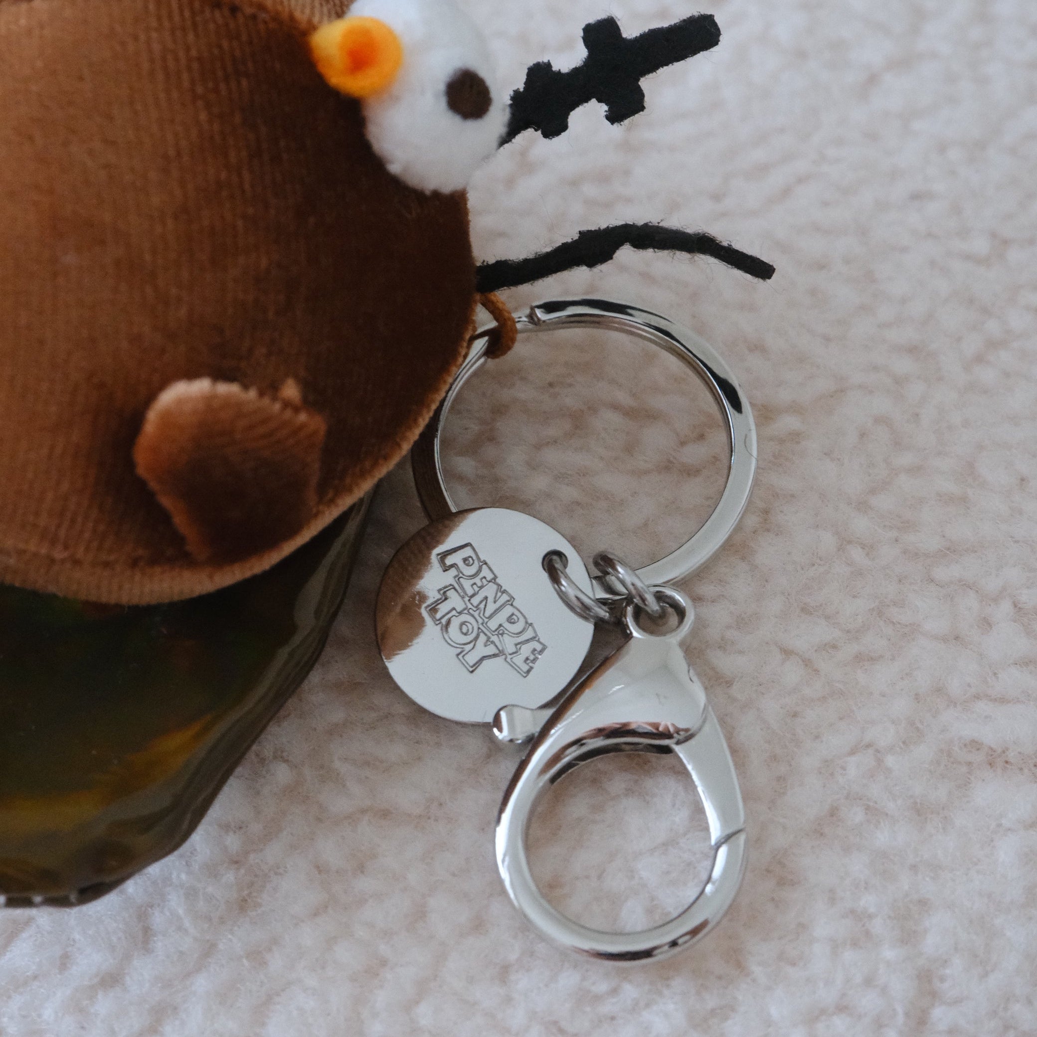 A small plush cockroach keychain with olive green wings, yellow feet, black antler-like details, and a metal keychain with a charm engraved with the text, PENPLE TOY. resting on a white textured surface against a plain light gray background.