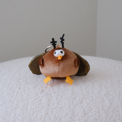 A small plush cockroach keychain with olive green wings, yellow feet, and black antler-like details, resting on a white textured surface against a plain light gray background.
