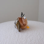 A side profile of a small plush cockroach keychain with olive green wings, yellow feet, and black antler-like details, resting on a white textured surface against a plain light gray background.
