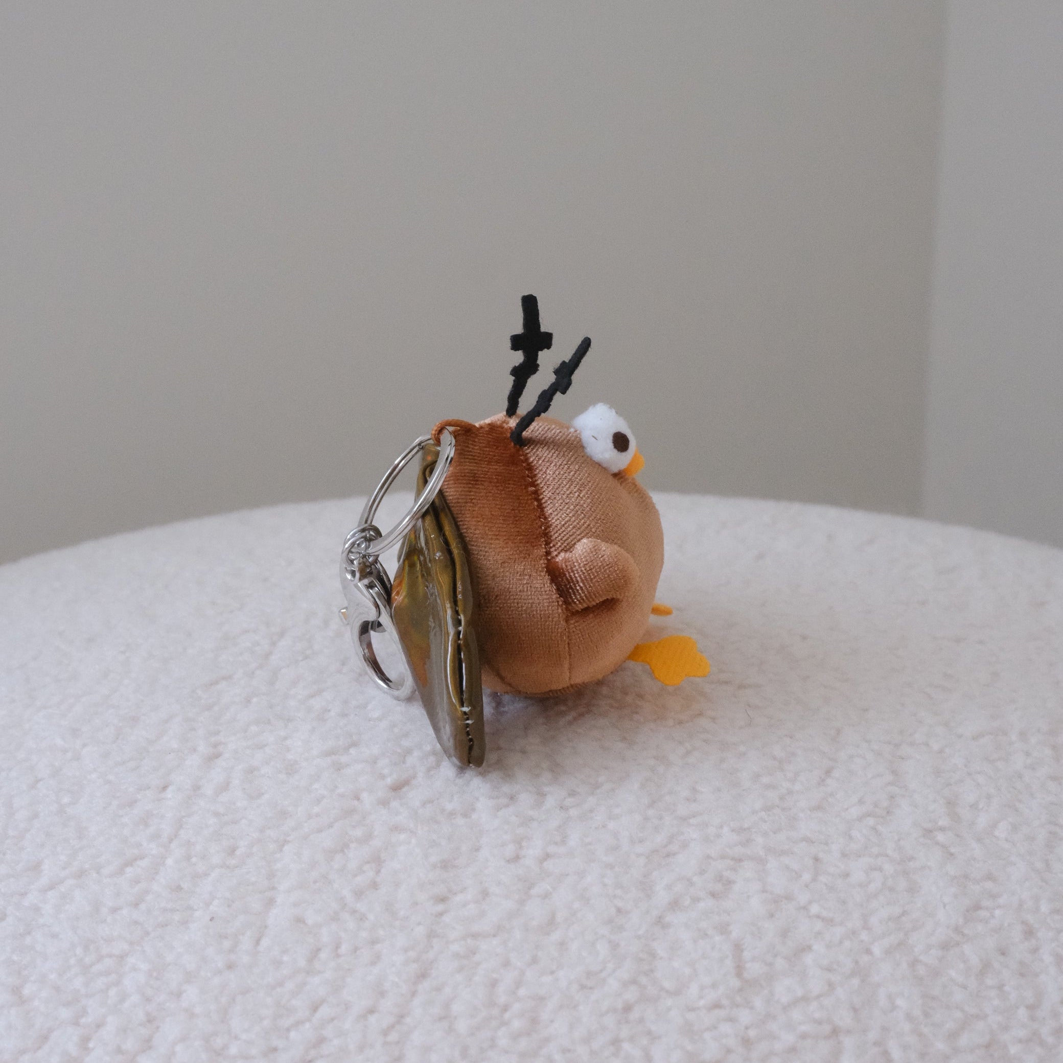 A side profile of a small plush cockroach keychain with olive green wings, yellow feet, and black antler-like details, resting on a white textured surface against a plain light gray background.