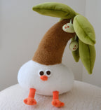 A cute plush toy shaped like a small coconut tree with a white round body, orange feet, a brown curved neck, green leaves, and a small orange beak, sitting on a textured white surface against a plain light gray background.