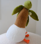 A side profile of a plush toy shaped like a coconut tree with a green leafy crown, brown neck, white body, and orange feet, sitting on a textured cream surface against a plain light background.