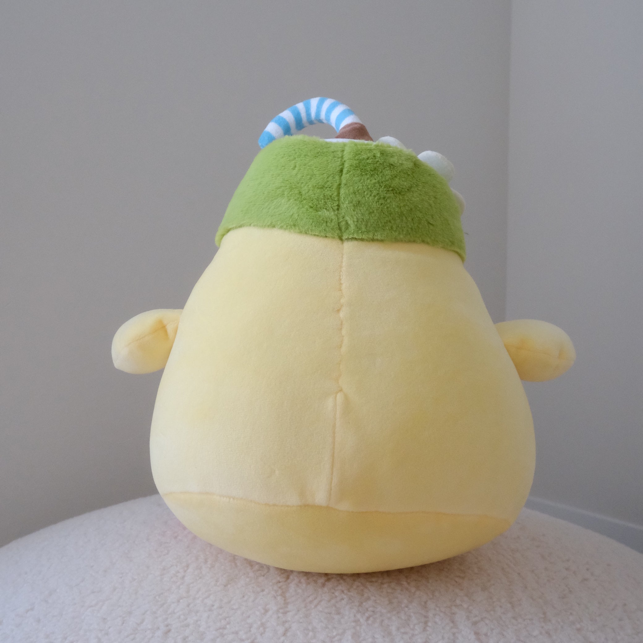 A yellow plush toy with a green coconut hat and a blue-and-white striped straw, photographed from the back against a plain indoor wall.