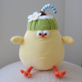 A yellow plush toy with orange feet and arms, wearing a green coconut hat with a white flower and a blue-and-white striped straw, sitting on a white textured surface against a plain light gray background.