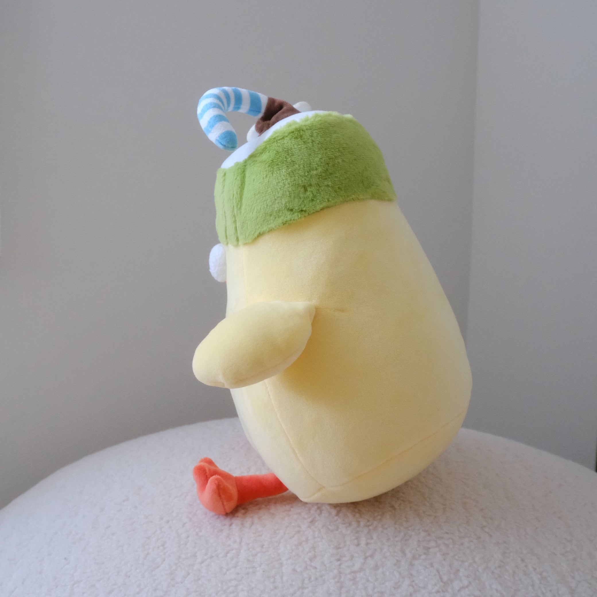 A side profile of a yellow plush toy with a green coconut hat and orange feet, sitting on a textured white surface against a plain gray wall.