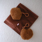 A brown suede-like rectangular pouch or case with two attached fuzzy brown pom-pom accessories and matching brown loops, laid flat on a white textured surface.