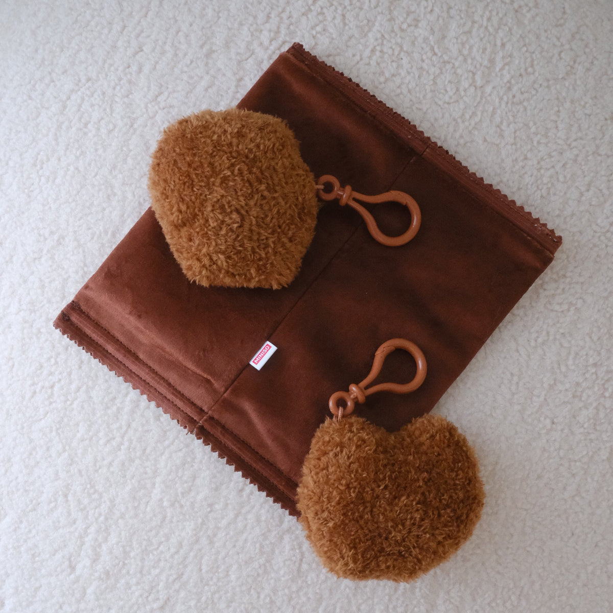 A brown suede-like rectangular pouch or case with two attached fuzzy brown pom-pom accessories and matching brown loops, laid flat on a white textured surface.