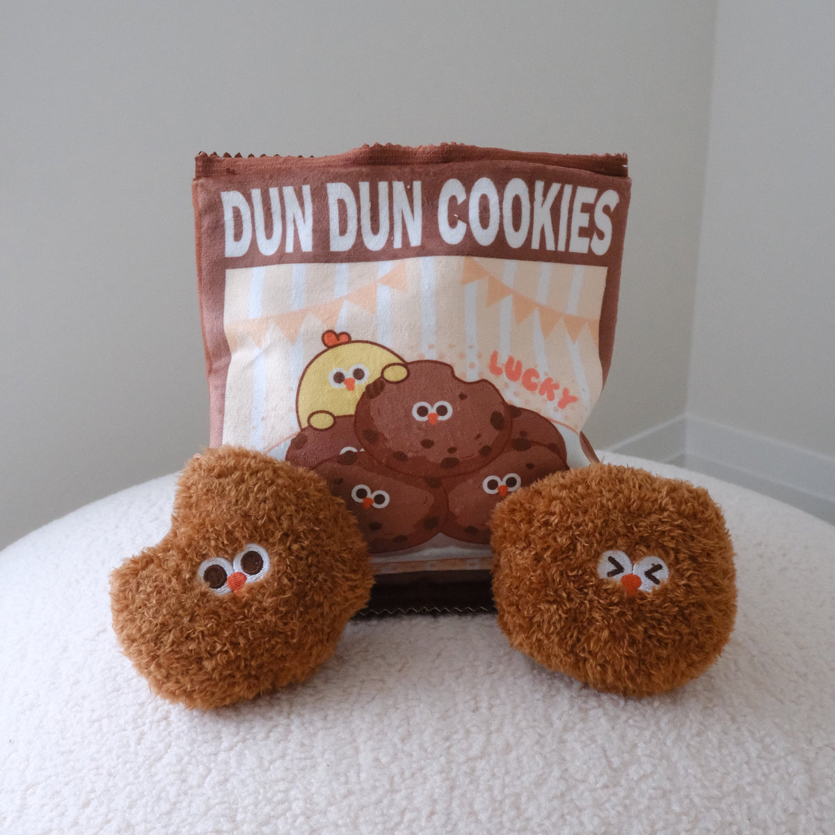 A bag of 'DUN DUN COOKIES' with two small brown plush character cookies placed in front on a white textured surface against a plain light gray wall.