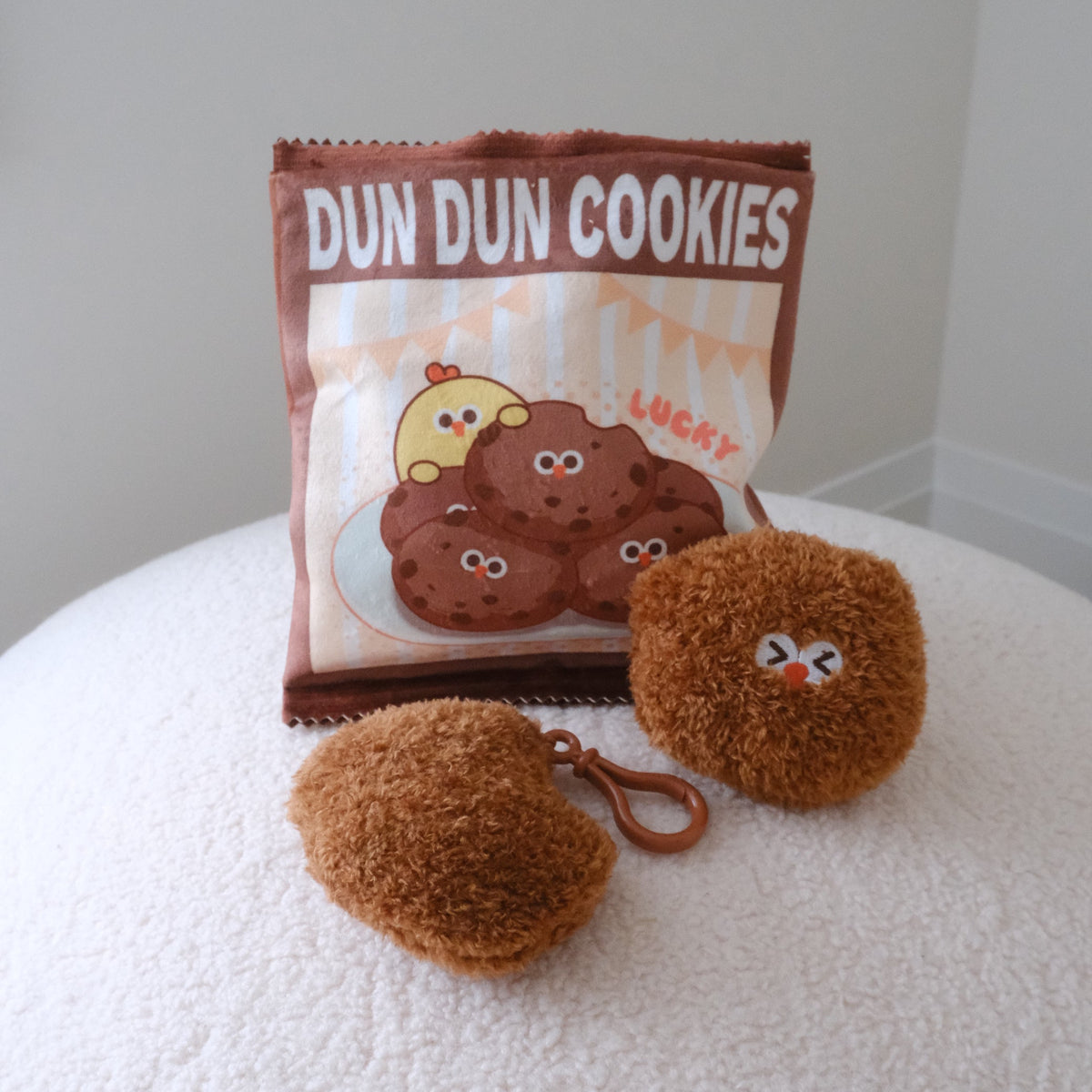 A brown plush cookie-shaped keychain and matching pouch labeled 'DUN DUN COOKIES' sit on a white textured surface against a light gray wall.
