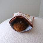 A brown plush toy with eyes peeking out from inside a suede brown bag that reads 'DUN DUN COOKIE', placed on a white textured surface against a plain light gray wall.