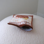 A small printed pouch or bag with a cartoon character and the text 'DUN DUN COOKIES' resting on a textured cream surface against a plain light gray wall.