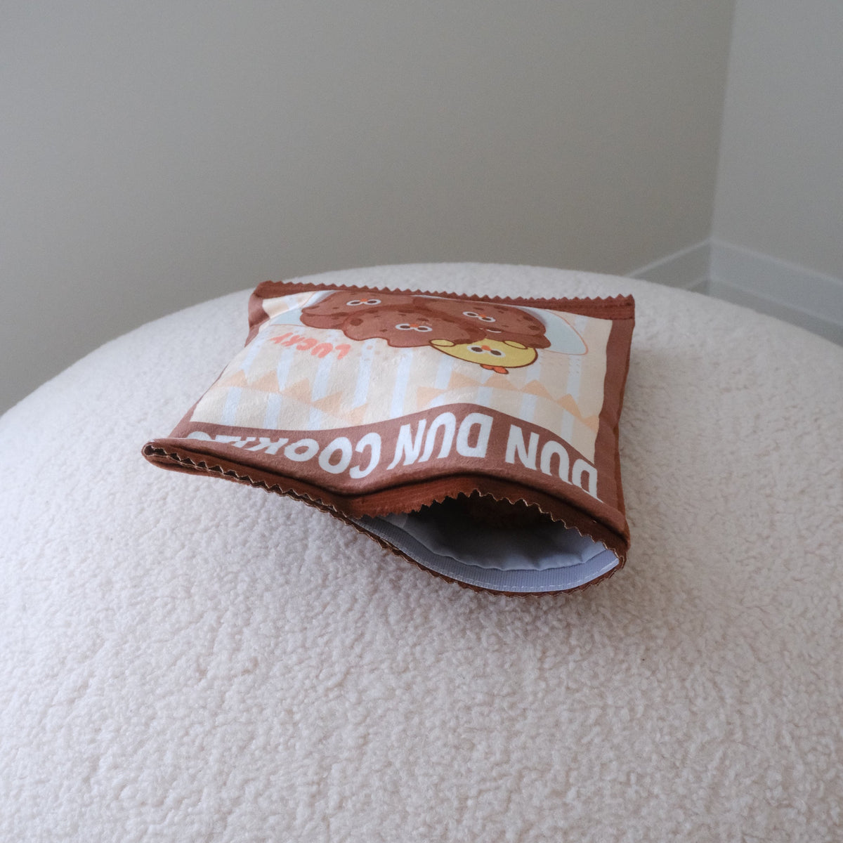 A small printed pouch or bag with a cartoon character and the text 'DUN DUN COOKIES' resting on a textured cream surface against a plain light gray wall.