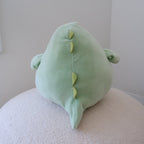 A light green plush dinosaur plush viewed from behind, resting on a textured cream-colored surface against a plain light gray wall.