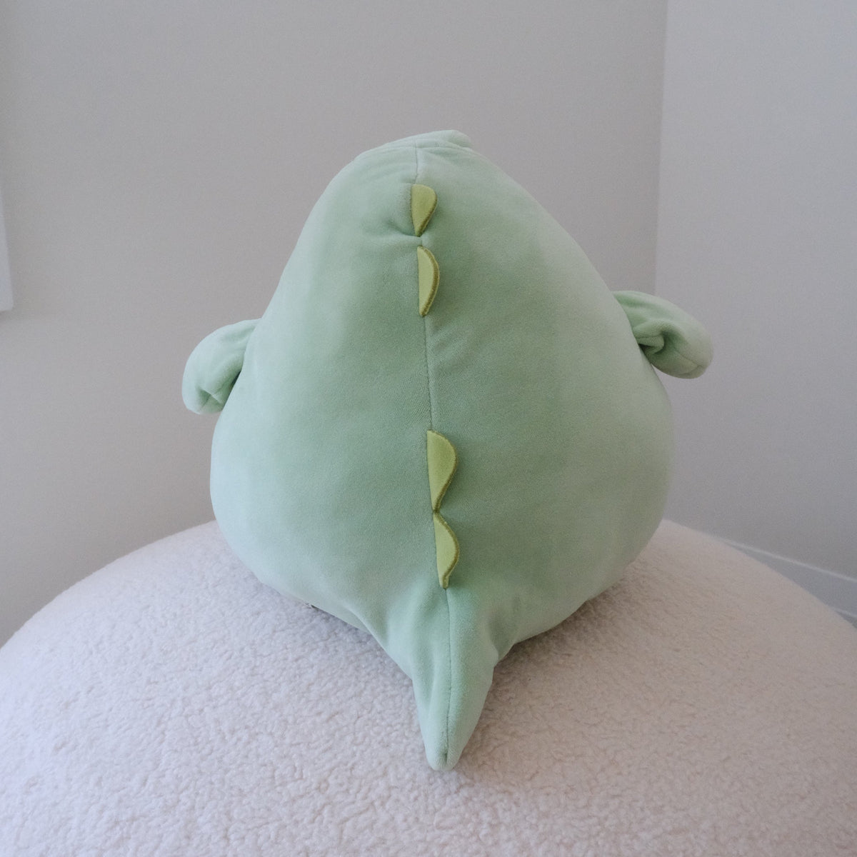 A light green plush dinosaur plush viewed from behind, resting on a textured cream-colored surface against a plain light gray wall.