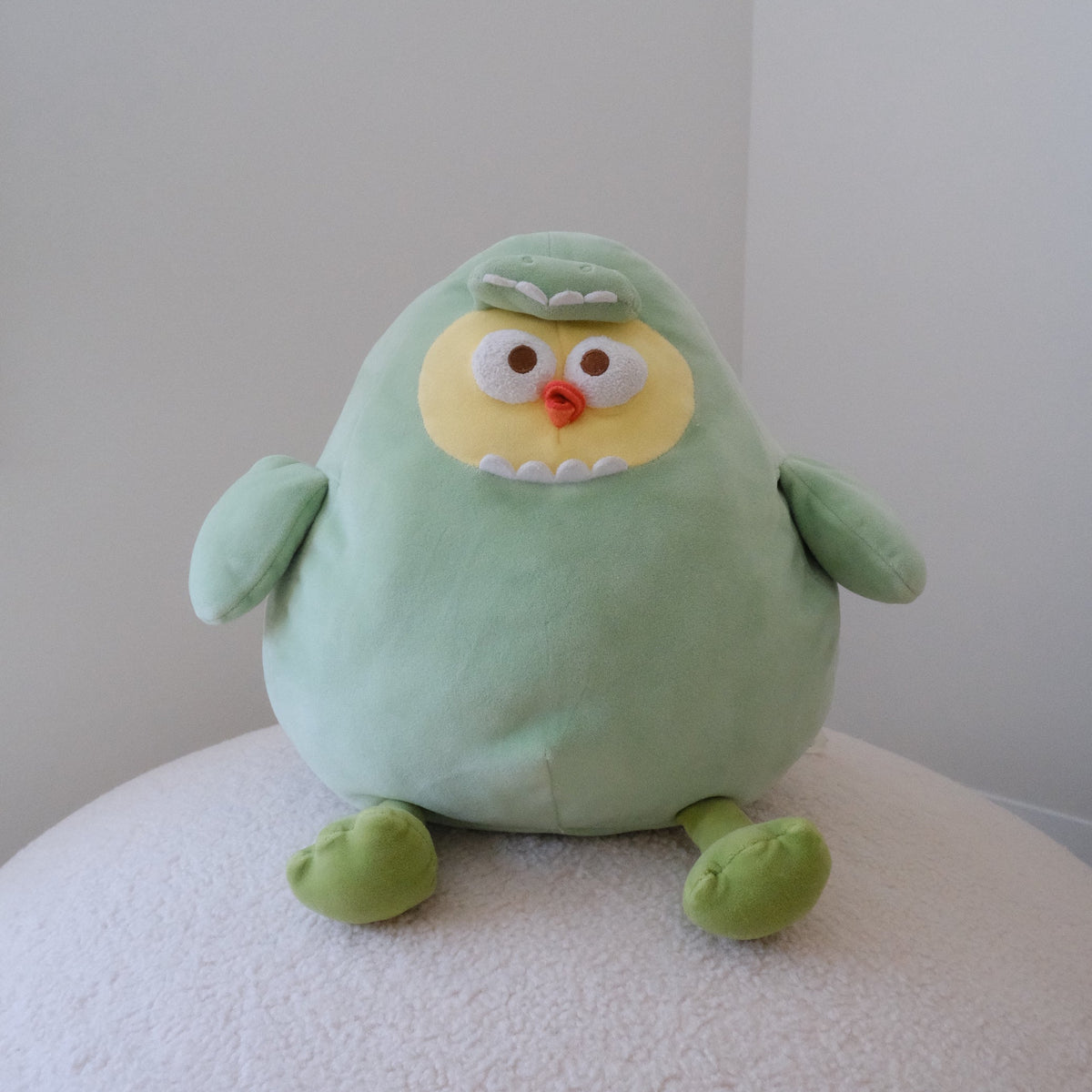A light green plush dinosaur toy with a yellow face, white eyes, and a small orange beak, sitting on a textured white surface against a plain light gray background.