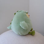 A light green plush dinosaur toy with small green spikes and a small yellow detail, sitting on a textured white surface against a plain light gray wall.