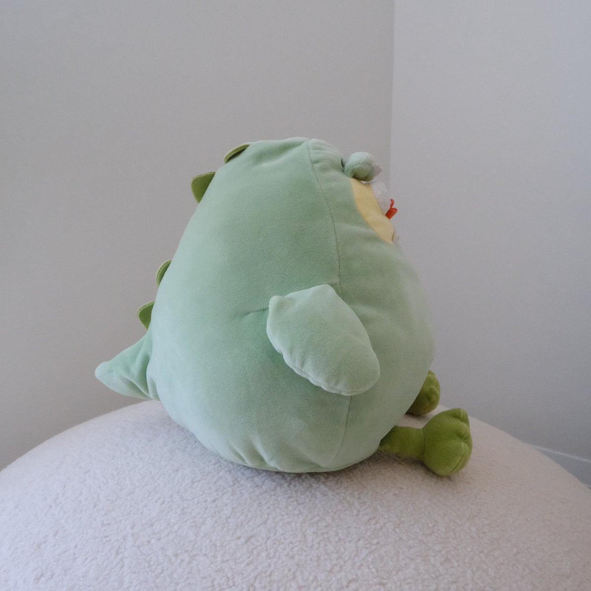 A light green plush dinosaur toy with small green spikes and a small yellow detail, sitting on a textured white surface against a plain light gray wall.