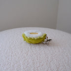 A small dundun chicken shaped like a quarter of a durian and a yellow pulp inside its shell, attached to a silver metal keyring, resting on a textured white surface against a plain beige wall.
