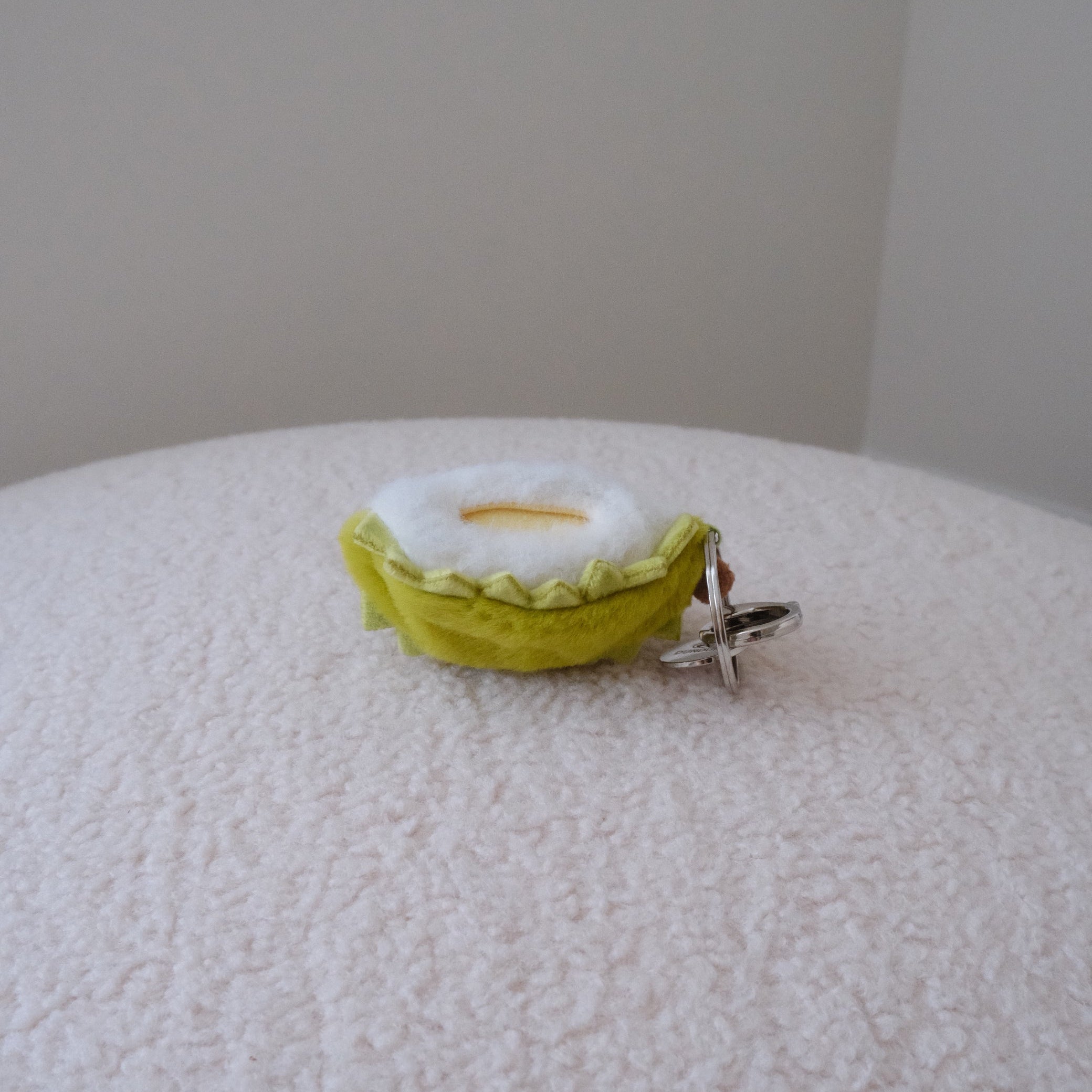 A small dundun chicken shaped like a quarter of a durian and a yellow pulp inside its shell, attached to a silver metal keyring, resting on a textured white surface against a plain beige wall.