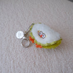 A small plush keychain shaped like a quarter slice of durian, featuring white plush fabric, light green ruffles, orange feet, and a silver metal keyring, resting on a textured cream-colored surface.