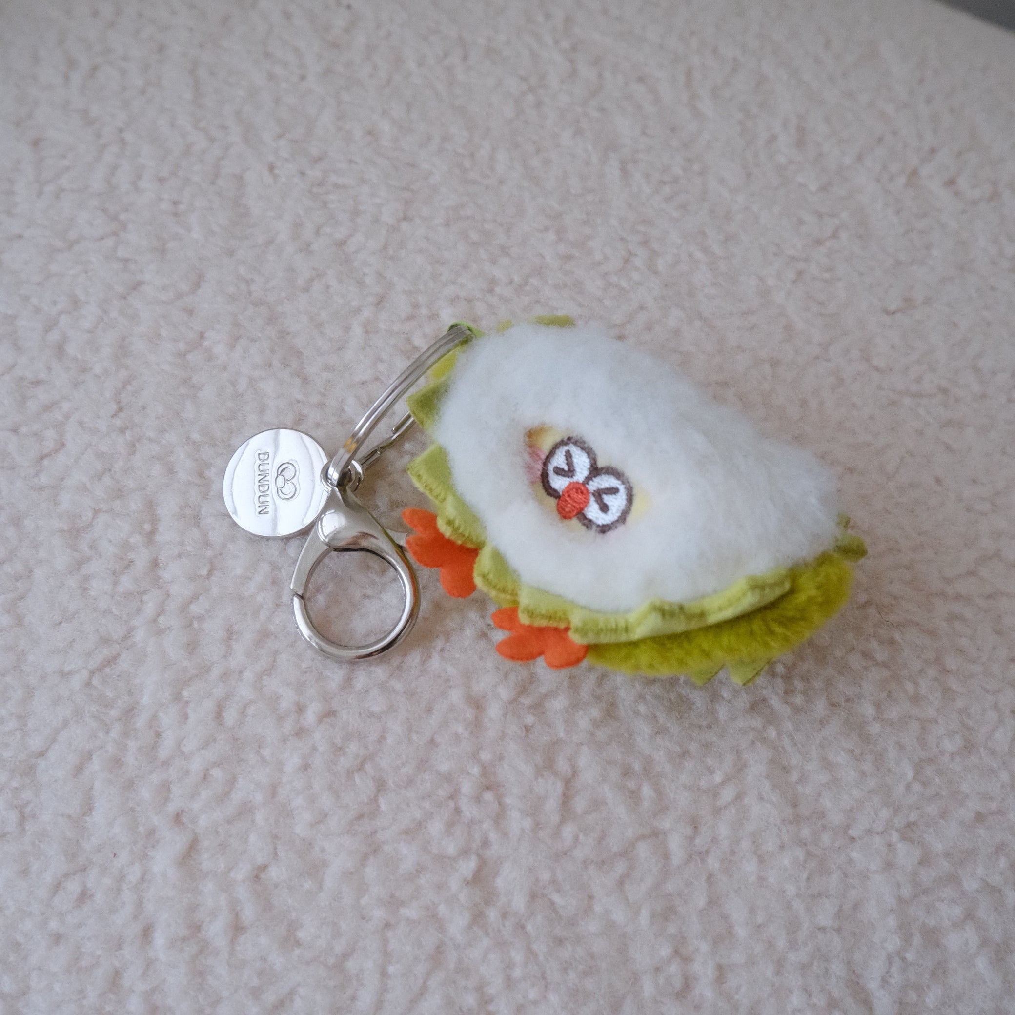 A small plush keychain shaped like a quarter slice of durian, featuring white plush fabric, light green ruffles, orange feet, and a silver metal keyring, resting on a textured cream-colored surface.