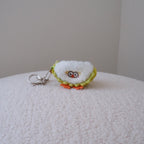 A small plush keychain shaped like a quarter of a durian with embroidered eyes and a yellow-green stitched border, attached to a silver metal key ring, resting on a textured cream surface against a plain light gray wall.