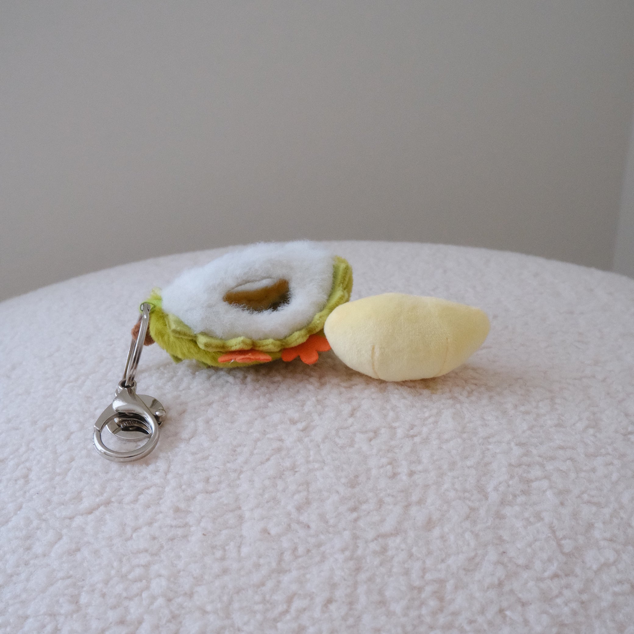 A small plush durian keychain resting on a textured white surface. The keychain features a yellow plush durian pulp attached to a green and white plush durian shell with a  silver metal clasp.