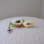 A small plush durian keychain resting on a textured white surface. The keychain features a yellow plush face with red eyes and pink cheeks, attached to a green and white plush durian shell with a  silver metal clasp.