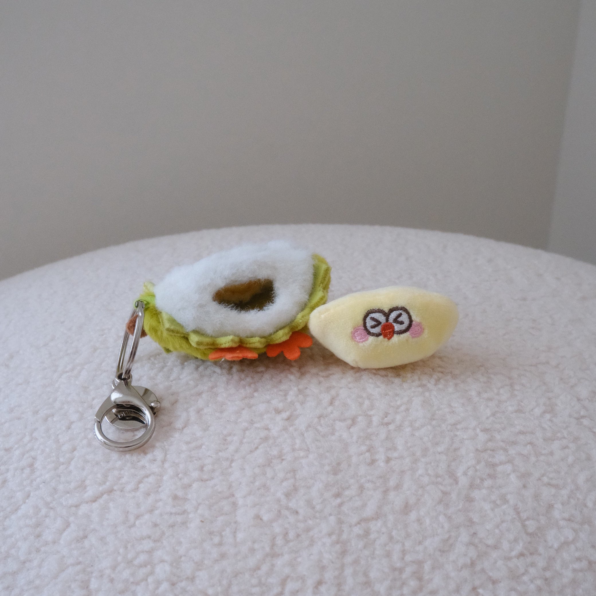 A small plush durian keychain resting on a textured white surface. The keychain features a yellow plush face with red eyes and pink cheeks, attached to a green and white plush durian shell with a  silver metal clasp.