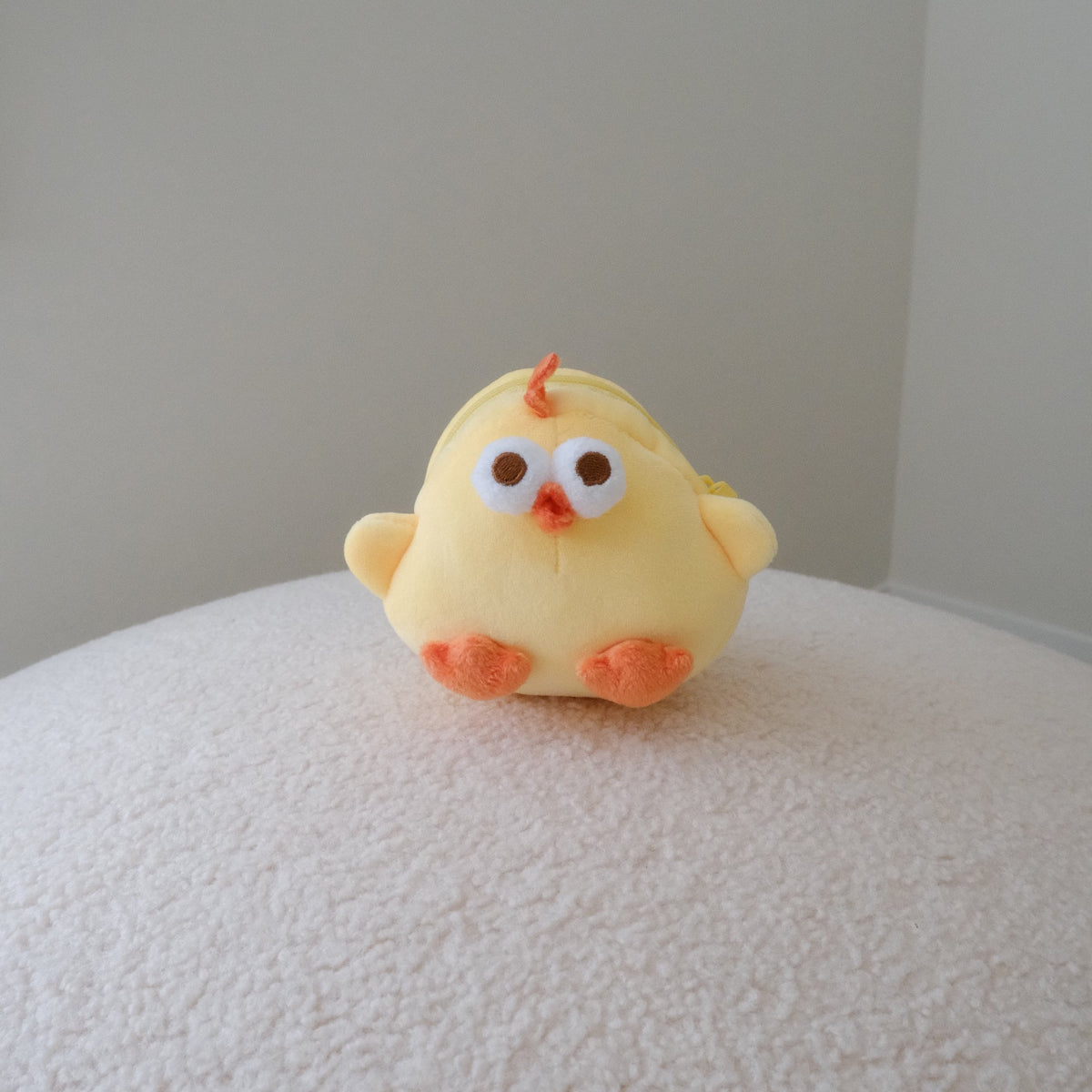 A small yellow plush chick pouch with orange feet and beak, sitting on a textured white surface against a plain light gray wall.