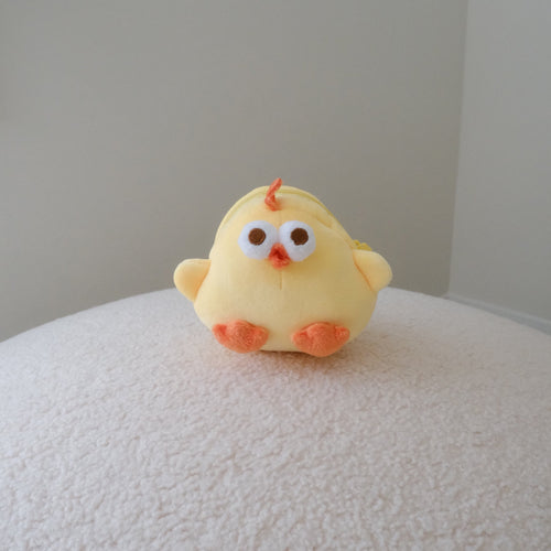A small yellow plush chick pouch with orange feet and beak, sitting on a textured white surface against a plain light gray wall.
