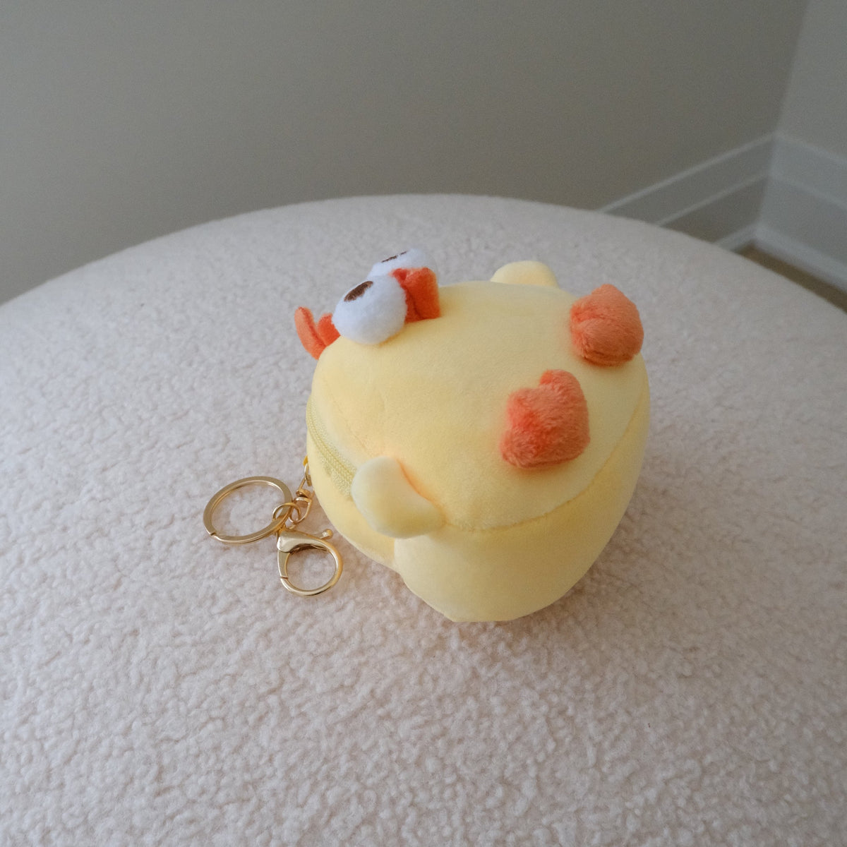 A small yellow plush character keychain or coin purse with orange heart-shaped details and a gold key ring, resting on a textured white surface against a neutral indoor background.