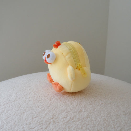A small yellow chick-shaped zippered pouch with orange feet and a white eye, resting on a textured white surface against a plain light gray background.