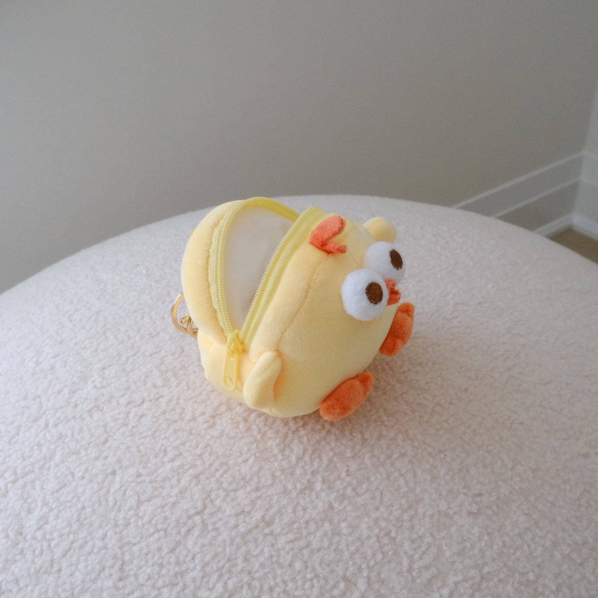 A small yellow chick-shaped zipper pouch with orange feet and a white face, resting on a textured white surface against a neutral beige wall.