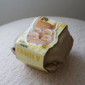 A brown paper-wrapped plush toy shaped like a family of four cartoon chicks, resting on a textured cream fabric surface against a neutral gray background.