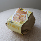 A brown paper-wrapped plush toy shaped like a family of four cartoon chicks, resting on a textured cream fabric surface against a neutral gray background.