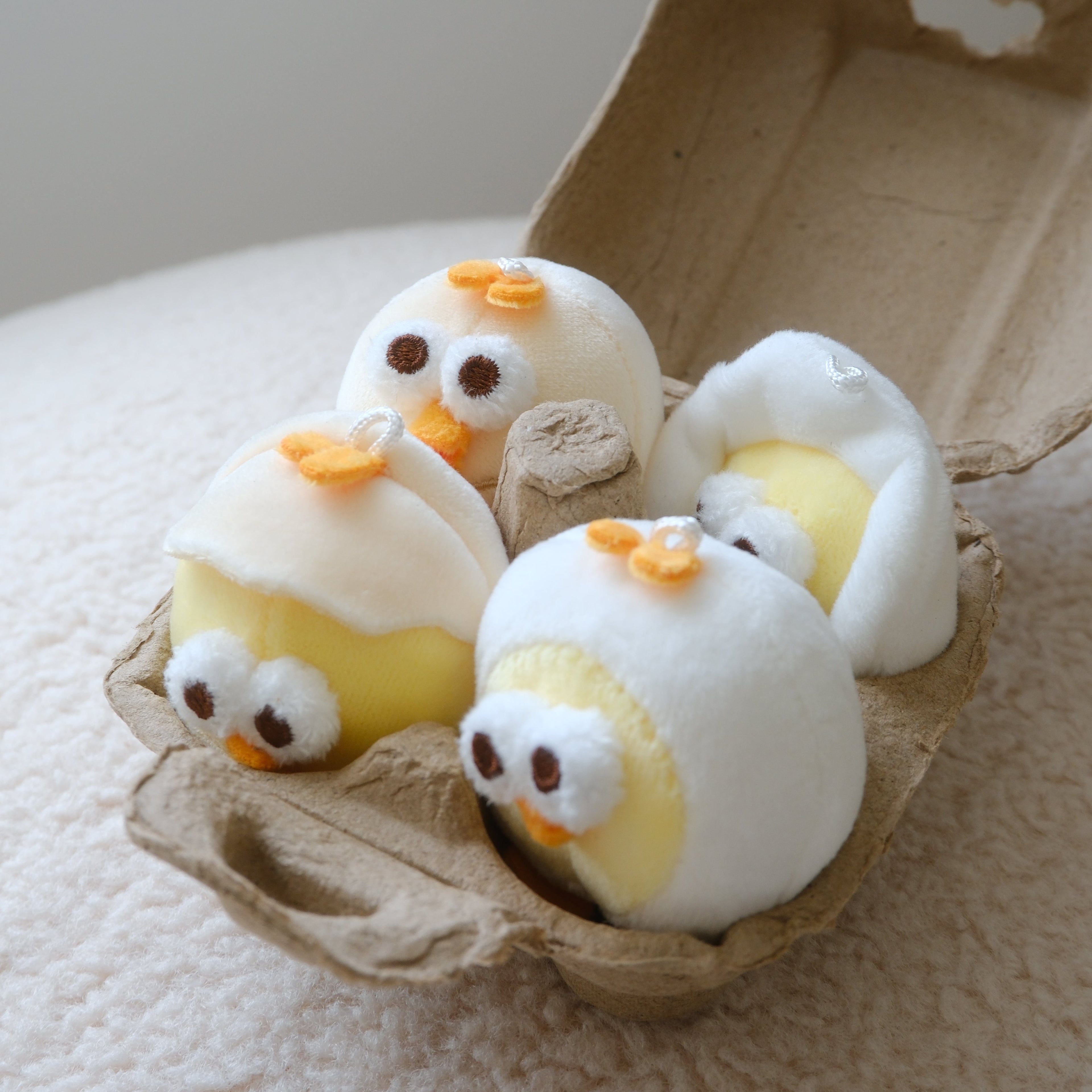 Four small plush chick toys with white bodies, yellow bellies, and orange beaks resting inside an open brown cardboard egg carton on a soft cream fabric surface.