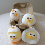 Four plush egg-shaped chick toys in white and yellow fabric, with orange beaks and feet, arranged around an open brown cardboard egg carton on a soft cream fabric surface.