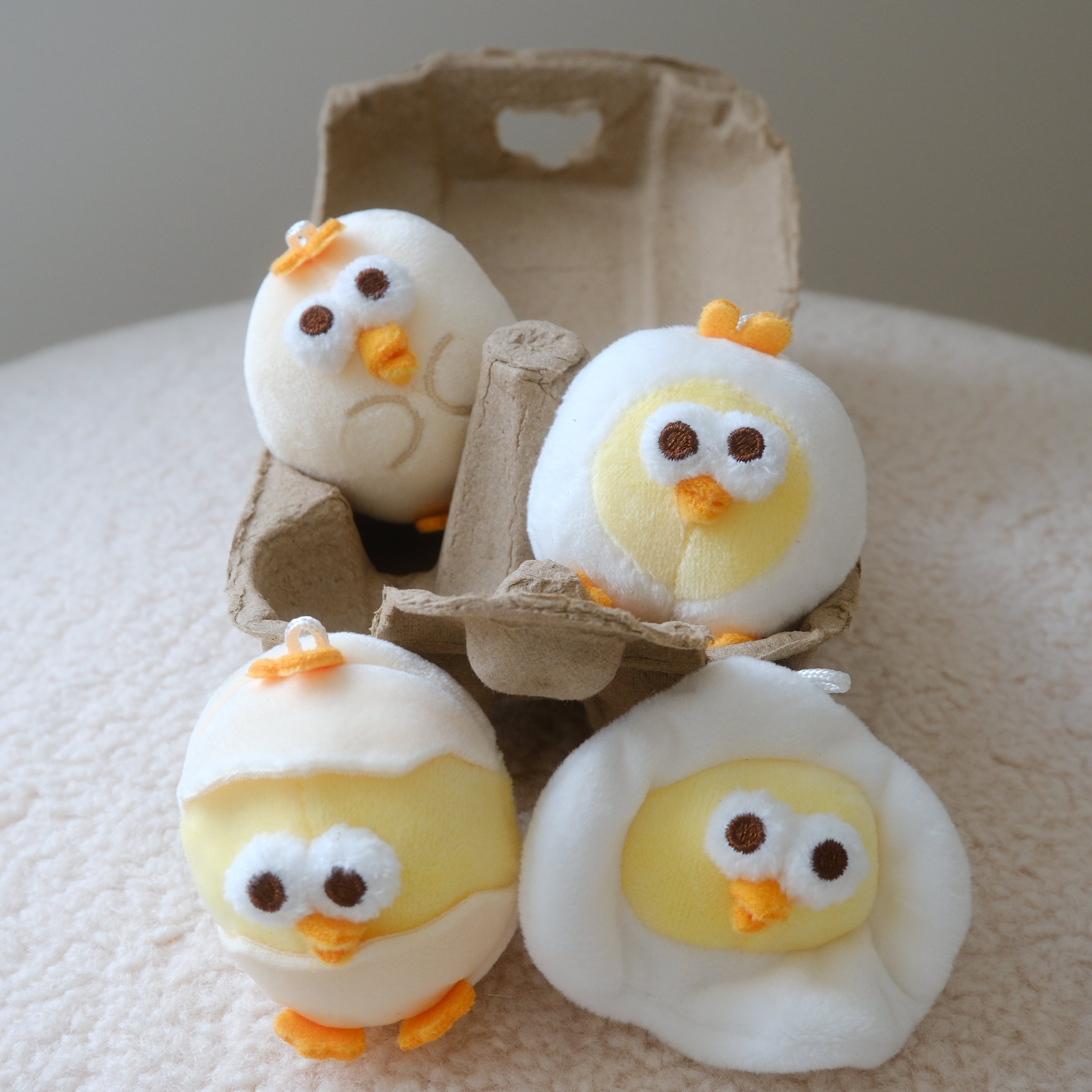 Four plush egg-shaped chick toys in white and yellow fabric, with orange beaks and feet, arranged around an open brown cardboard egg carton on a soft cream fabric surface.