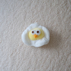 A small plush toy shaped like a fried egg with a yellow chick face peeking out from white fluffy edges, placed on a textured cream carpet.