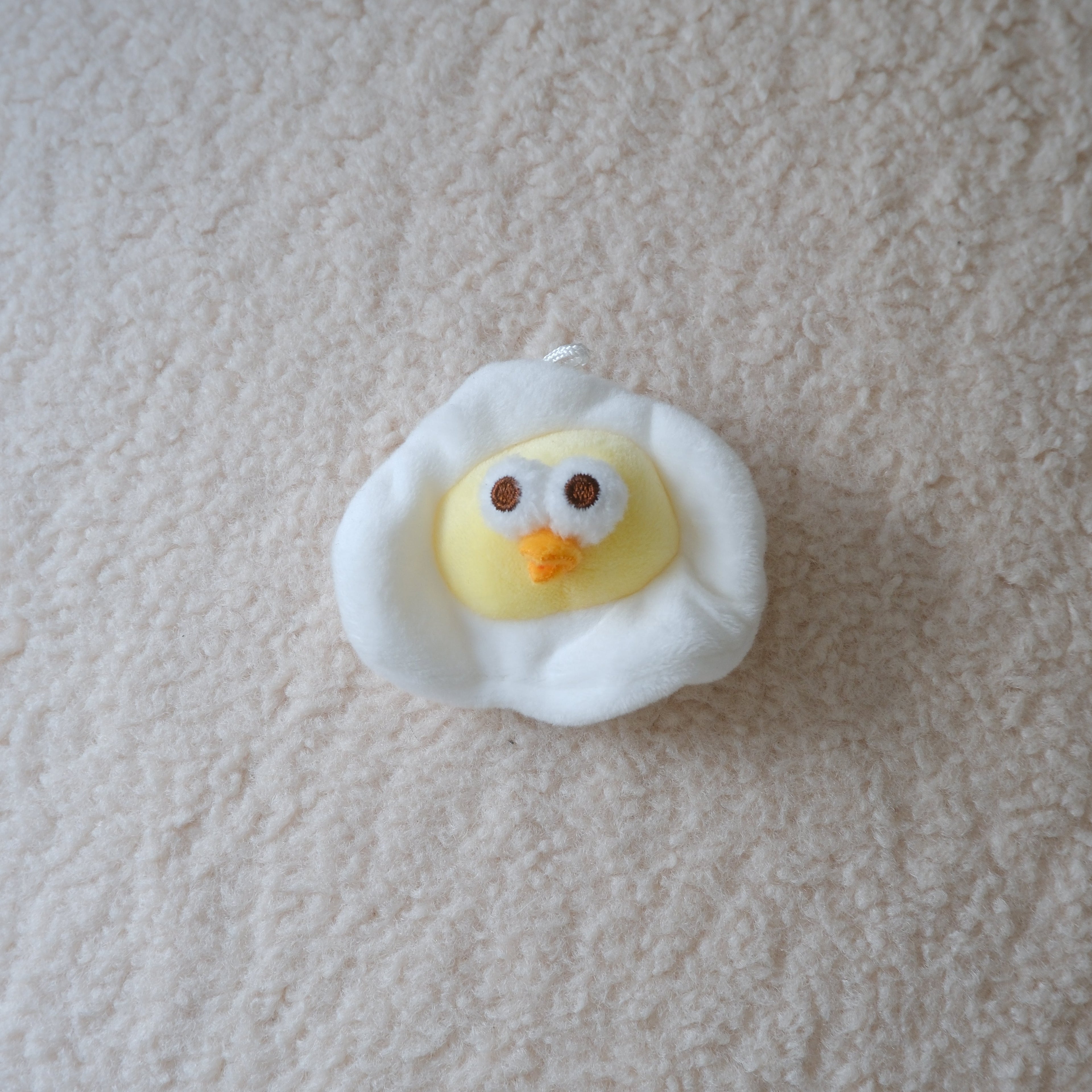A small plush toy shaped like a fried egg with a yellow chick face peeking out from white fluffy edges, placed on a textured cream carpet.